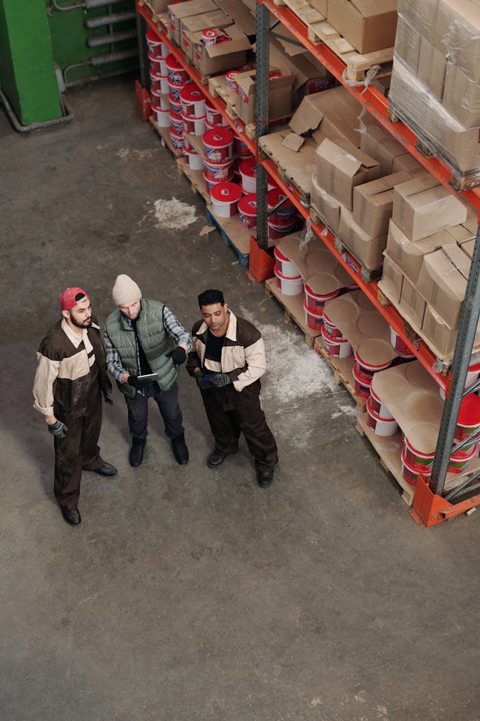 about-us Three workers in a warehouse discussing inventory management near shelves with boxes and paint cans.
